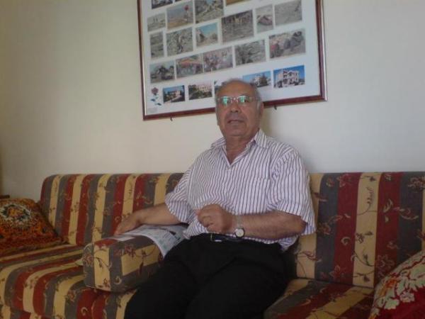 الأستاذ عبد الأمير علي مهنا في منزله وخلفه جدارية لصور الإعتداءات الصهيونية